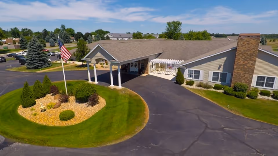 Exterior view of a single-story assisted living facility with a covered entrance, a circular driveway, and a landscaped roundabout featuring an American flag and various shrubs. The building has beige siding, white trim, and a brick chimney, surrounded by green lawns and trees under a clear blue sky.