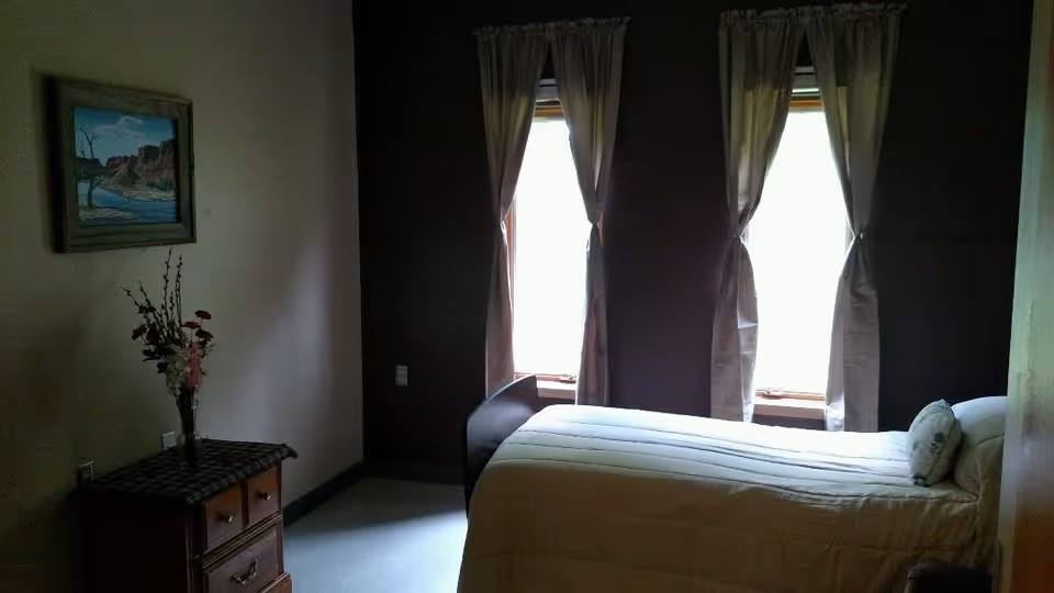A small bedroom with a single bed covered in a beige comforter and a pillow. Two windows with light-colored curtains allow natural light to enter the room. A wooden nightstand with a checkered cloth and a vase of flowers is positioned next to the bed. A framed landscape painting hangs on the wall above the nightstand.