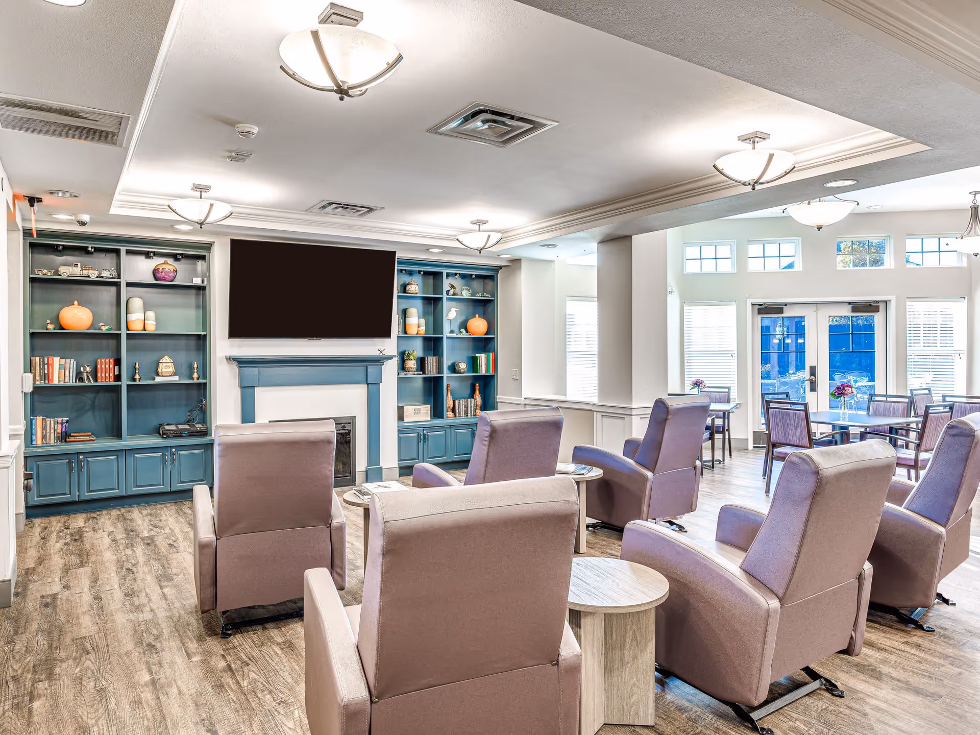 A bright and spacious common living area in a senior living facility featuring six comfortable armchairs arranged around small round tables. The room has wood flooring, a blue built-in bookshelf with decorative items and books, a fireplace with a large flat-screen TV mounted above it, and large windows and glass doors letting in natural light. In the background, there are dining tables and chairs near the windows.