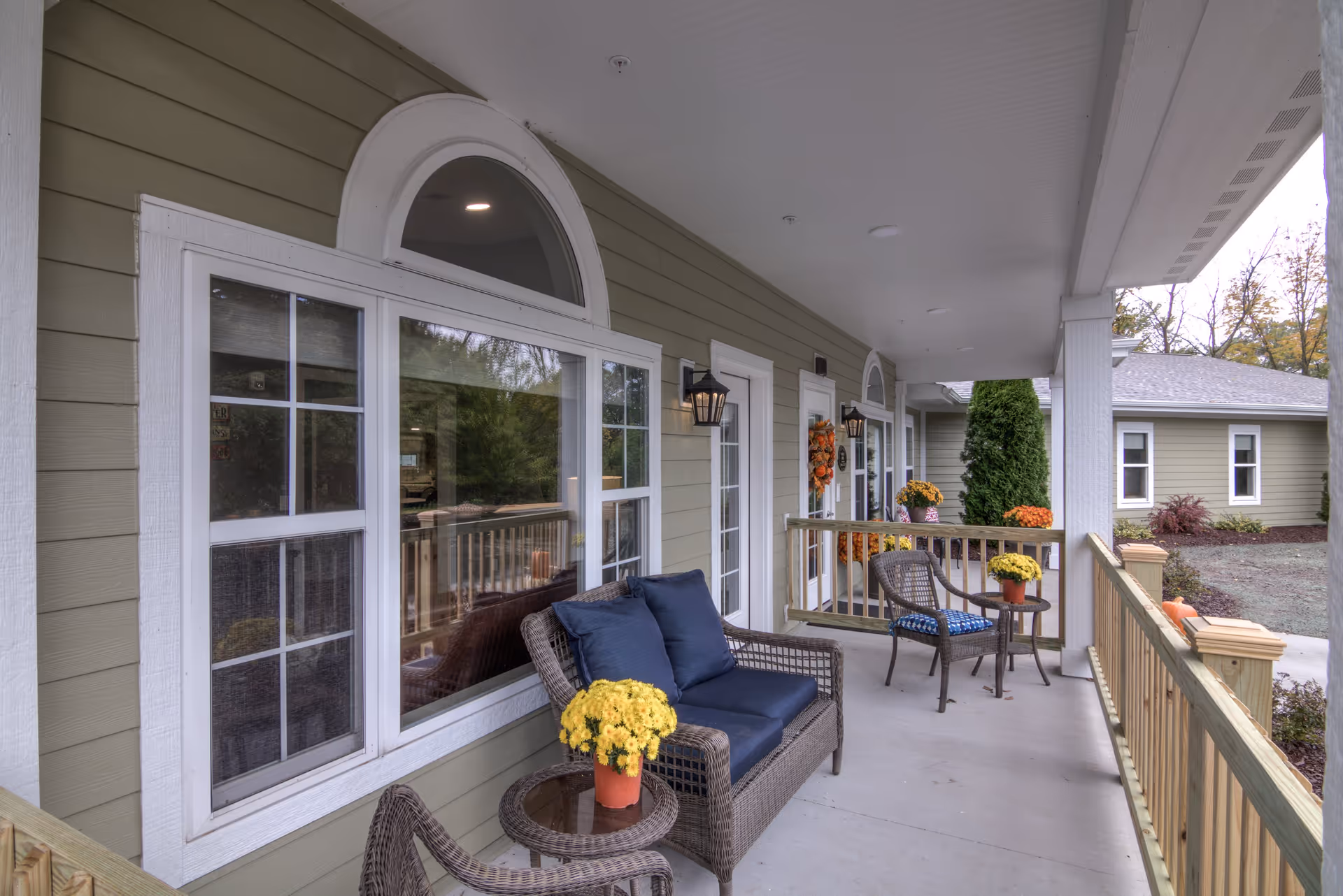 Covered porch area at Dimensions Living Sheboygan with wicker seating including a loveseat with blue cushions and two chairs, small round tables with yellow potted flowers, and fall decorations on the doors. The porch has a wooden railing and overlooks a landscaped yard with shrubs and trees.