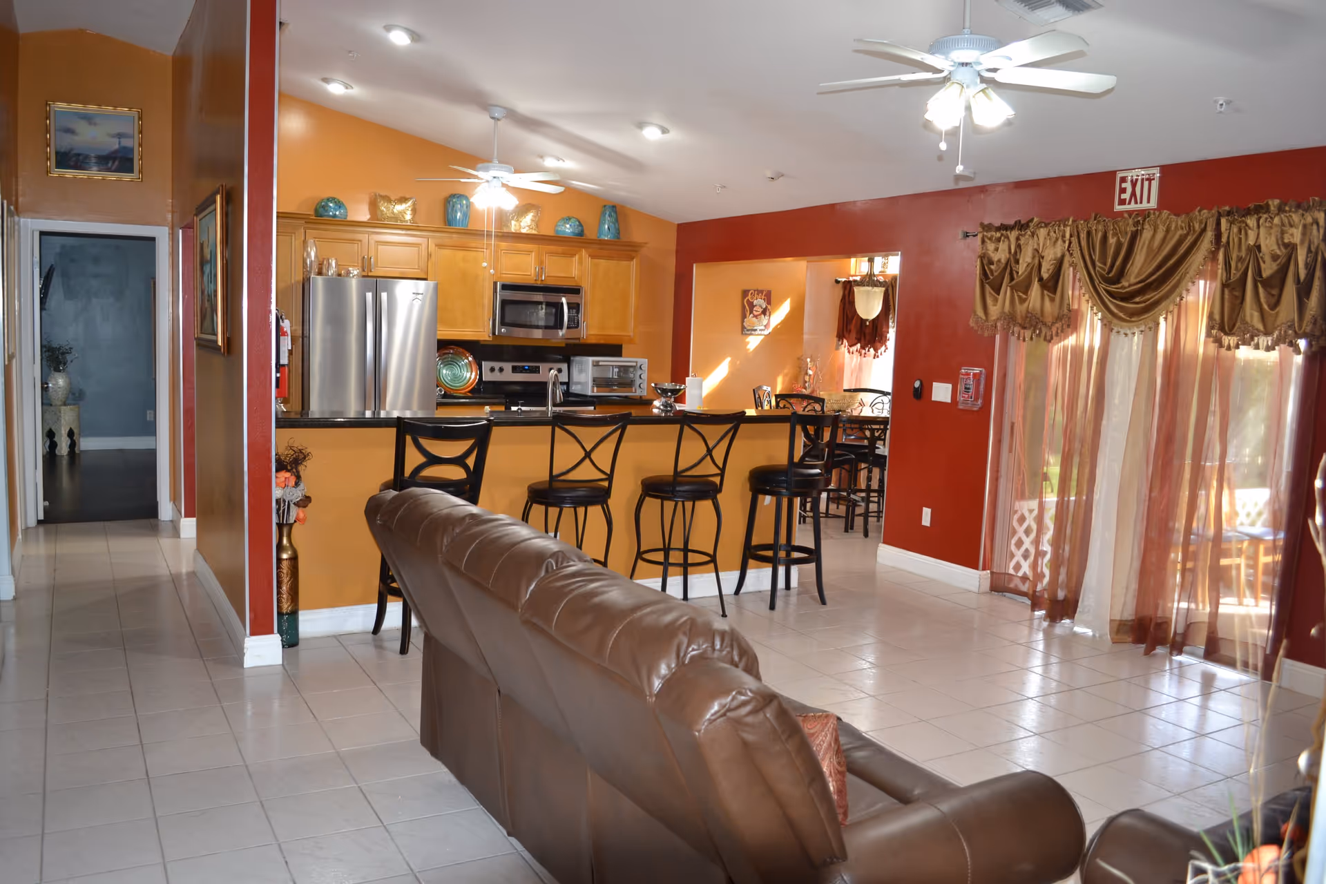 Interior view of a senior living facility showing a living room area with a brown leather couch facing a kitchen with a breakfast bar and four black bar stools. The kitchen has wooden cabinets, a stainless steel refrigerator, microwave, and oven. The walls are painted in warm tones of orange and red, with decorative vases on top of the cabinets. There is a ceiling fan with lights above the kitchen and another in the living room. To the right, sheer brown curtains cover a sliding glass door with an exit sign above it. A hallway leads to another room on the left side of the image.