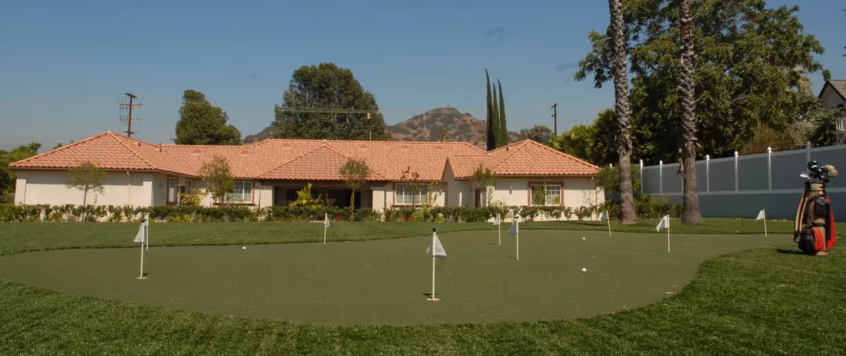 A single-story building with a red tile roof behind a putting green with several small flags and golf balls. There are trees and a mountain in the background under a clear blue sky.