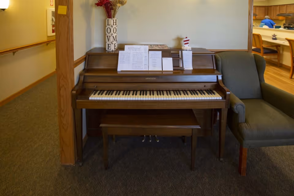 An upright piano with sheet music sits in a communal interior area beside an upholstered armchair and nearby dining space.