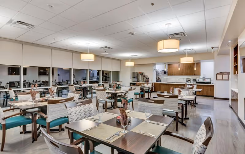 Spacious dining room with multiple set tables and chairs, pendant lights, and a serving counter and kitchen area in the back.