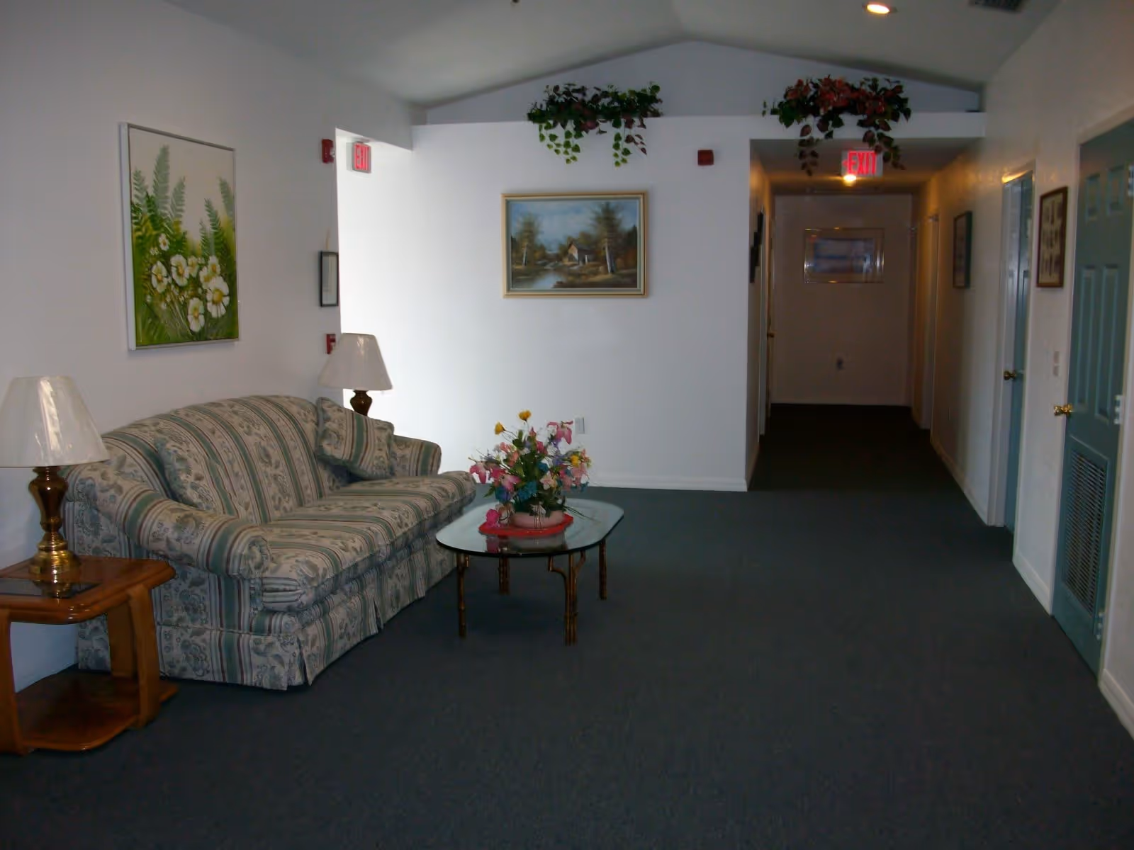 A hallway area with a patterned sofa, two table lamps on wooden side tables, a glass coffee table with a floral arrangement, and framed paintings on the walls. The hallway has carpeted flooring and white walls with some greenery decorations near the ceiling and exit signs.