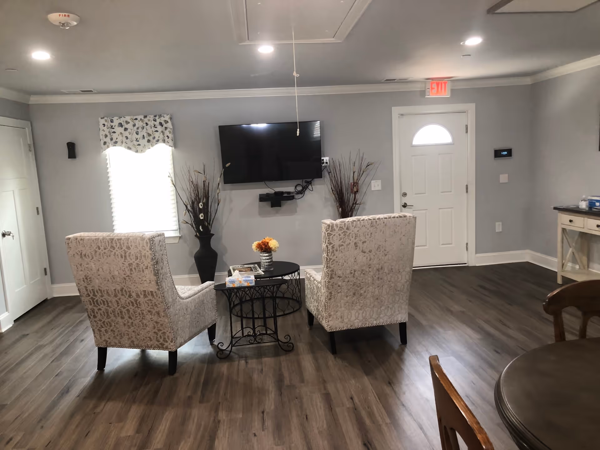 A cozy senior living common area with two patterned armchairs facing a wall-mounted flat screen TV. A small round table with a flower vase and magazines is placed between the chairs. The room has wood flooring, light gray walls, a window with a floral valance, and a white door with an exit sign above it. Decorative tall vases with dried plants are positioned on either side of the TV.