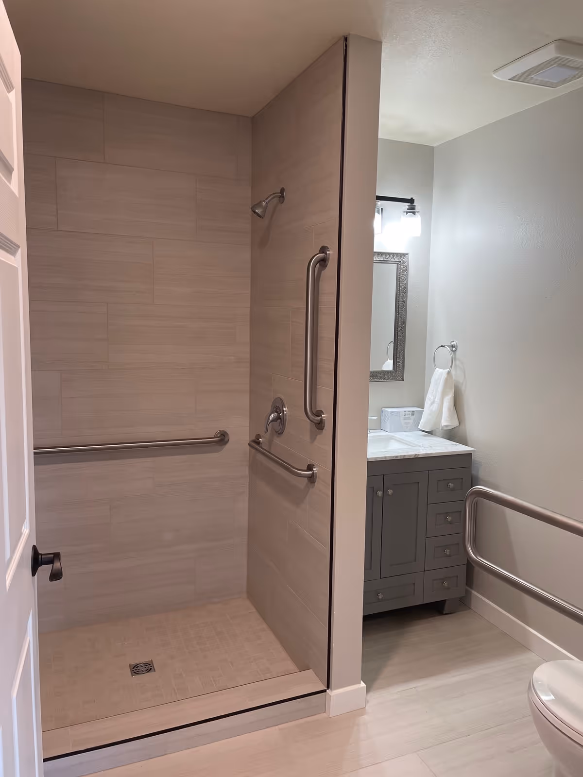 A modern bathroom featuring a walk-in shower with beige tiles and multiple stainless steel grab bars. The shower has a wall-mounted showerhead and a drain on the tiled floor. Adjacent to the shower is a gray vanity with a marble countertop, a mirror with a decorative frame, a towel ring holding a white towel, and a toilet with a grab bar next to it.