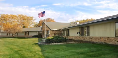 Single-story senior living facility exterior with brick accents, manicured lawn, and an American flag.