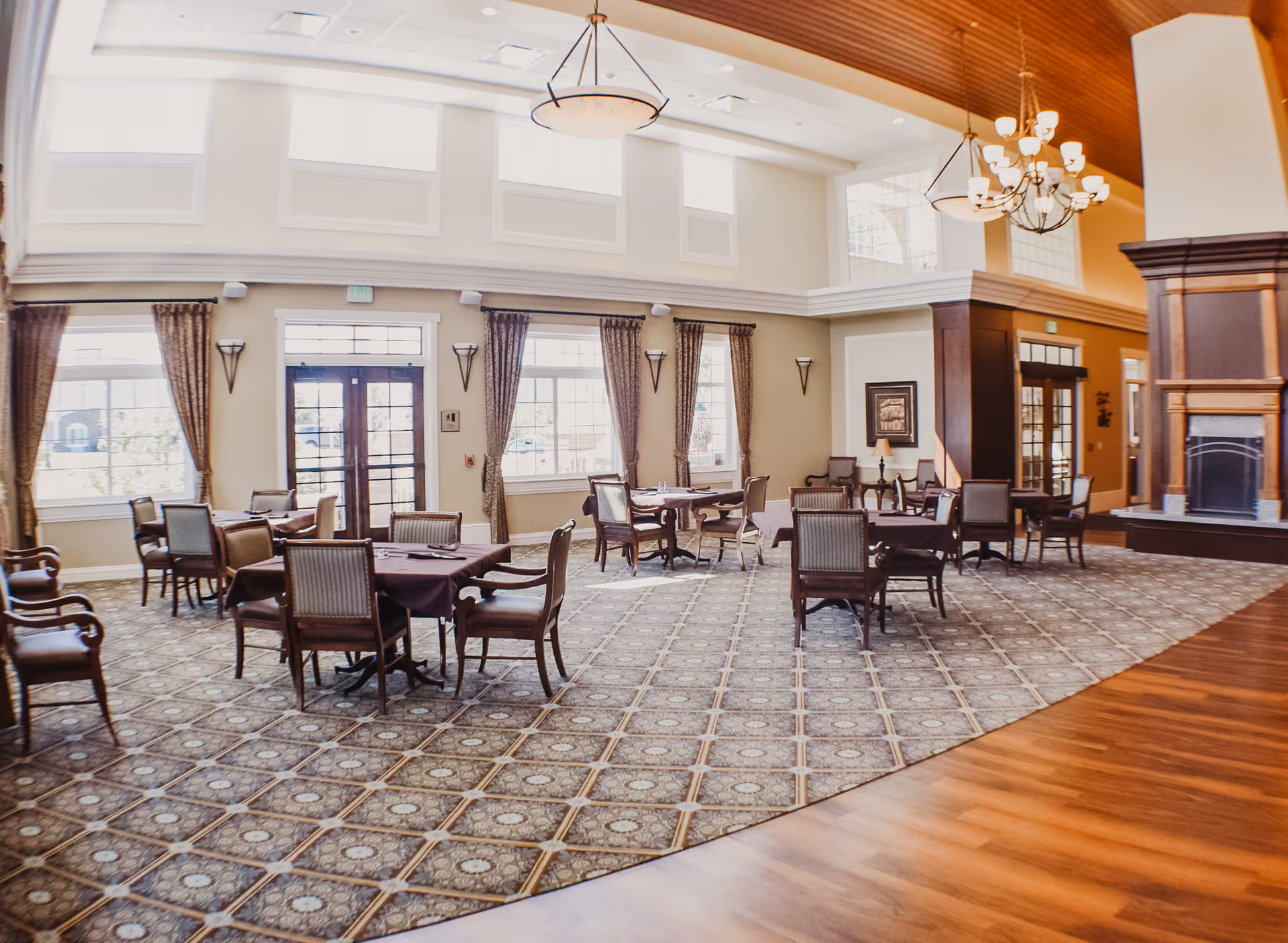 A spacious, well-lit dining area with multiple tables covered in dark tablecloths and surrounded by wooden chairs. Large windows with curtains allow natural light to fill the room. The room features high ceilings with chandeliers and a fireplace on the right side. The floor is a combination of patterned carpet and wood.