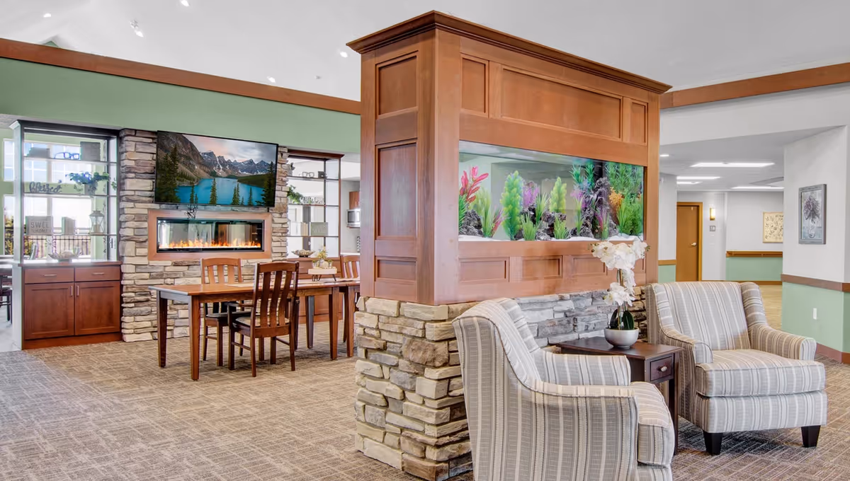 A cozy senior living facility common area featuring two striped armchairs with a small table and a potted orchid between them. A large wooden and stone partition with a built-in aquarium separates the seating area from a dining space with a wooden table and chairs. The dining area also has a stone fireplace and a mounted flat-screen TV above it. The room has carpeted floors, green and white walls, and built-in shelving with decorative items.