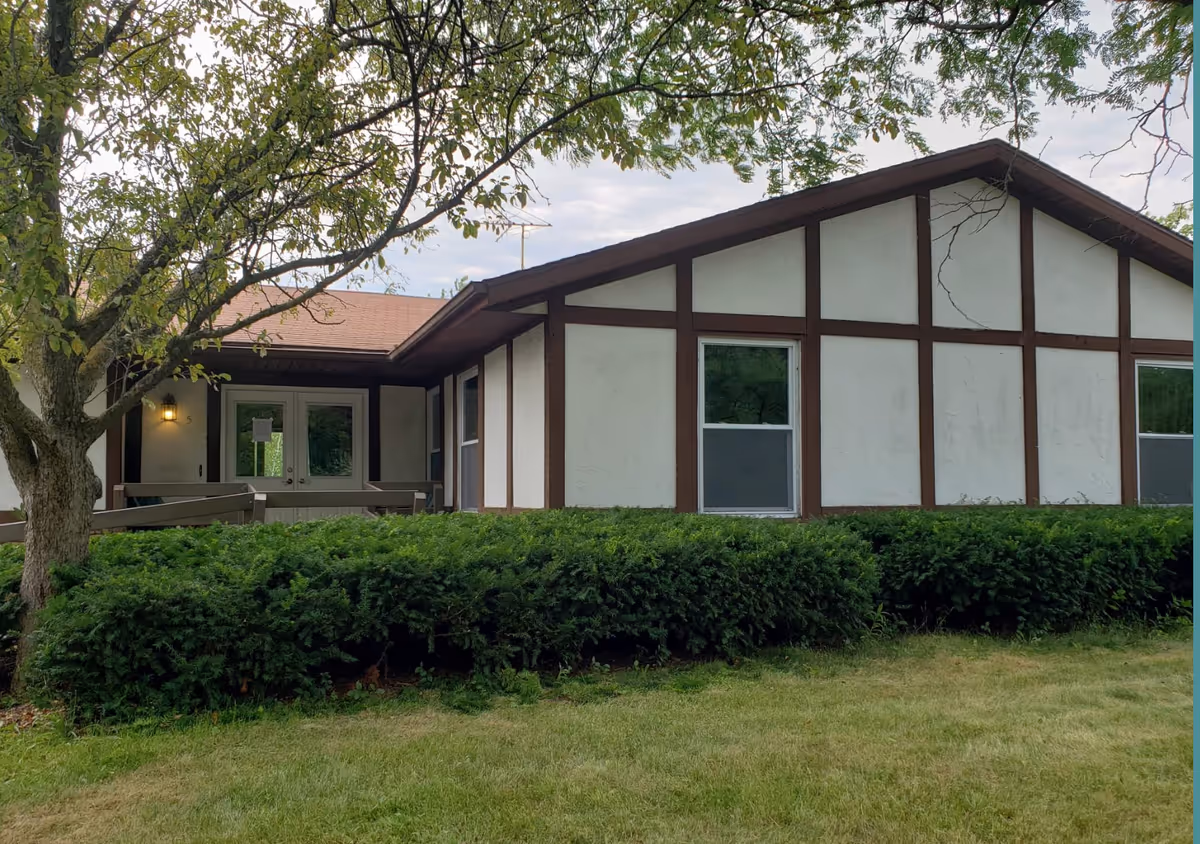 Exterior view of a single-story building with white walls and brown wooden trim. The building has a sloped roof and two windows visible on the right side. There is a small porch area with double doors and a ramp leading up to it. In front of the building, there are green bushes and a tree with green leaves. The sky is partly cloudy.
