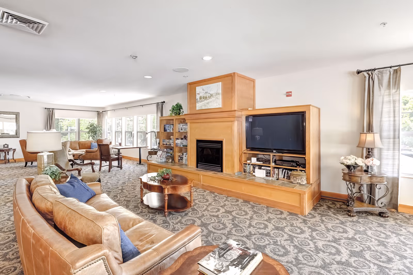 A spacious and well-lit living room area in an assisted living facility featuring a large tan leather sofa with blue pillows, a wooden coffee table with decorative items, a built-in wooden entertainment center with a flat-screen TV and fireplace, several armchairs, large windows with curtains allowing natural light, and a patterned carpeted floor.
