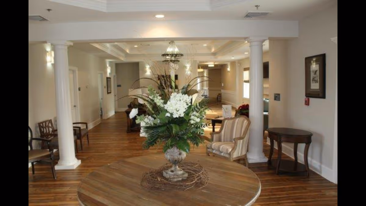 Interior view of a senior living facility hallway with wooden flooring, white columns, and a round wooden table in the foreground holding a large floral arrangement. There are chairs and small tables along the walls, soft lighting, and framed artwork on the walls.