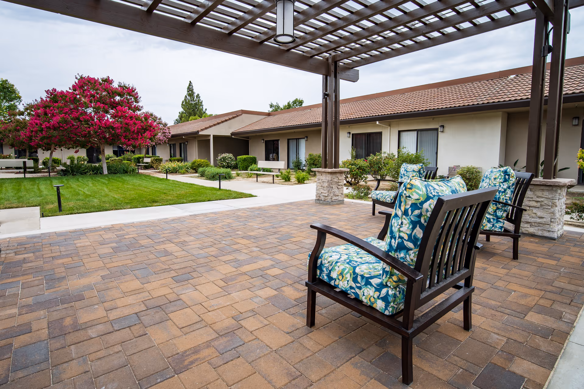 Patio courtyard at an assisted living facility with cushioned chairs under a pergola, paved patio, lawn and single-story buildings in the background.