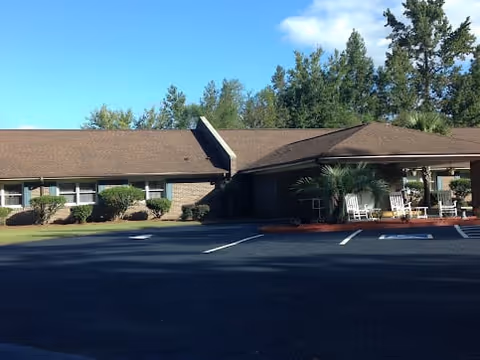 Exterior view of a single-story assisted living facility building with a brown roof and brick walls. The entrance has a covered porch with white rocking chairs. There are bushes and trees around the building and a clear blue sky above.
