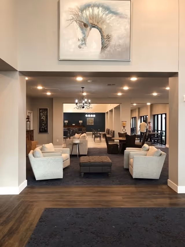 A spacious and well-lit common area in a senior living facility featuring four beige armchairs arranged around a central tufted ottoman on a dark carpet. In the background, there are multiple seating areas with tables and chairs, a chandelier, and large windows letting in natural light. A large abstract painting is mounted on the wall above the entrance to the room.