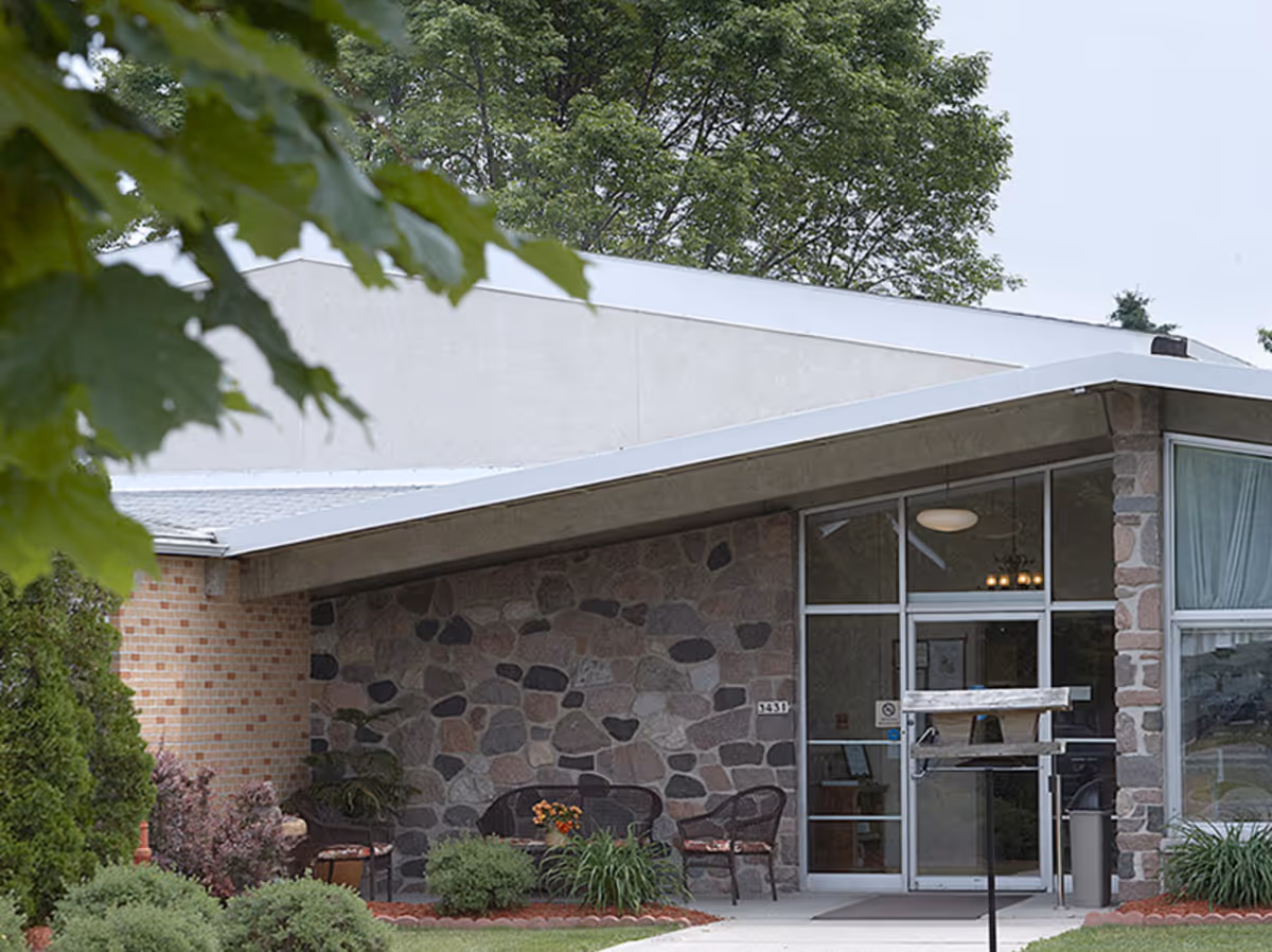Entrance of a building with a stone and brick exterior, featuring a glass door and large windows. There are outdoor chairs and a small table with flowers near the entrance, surrounded by greenery and trees.