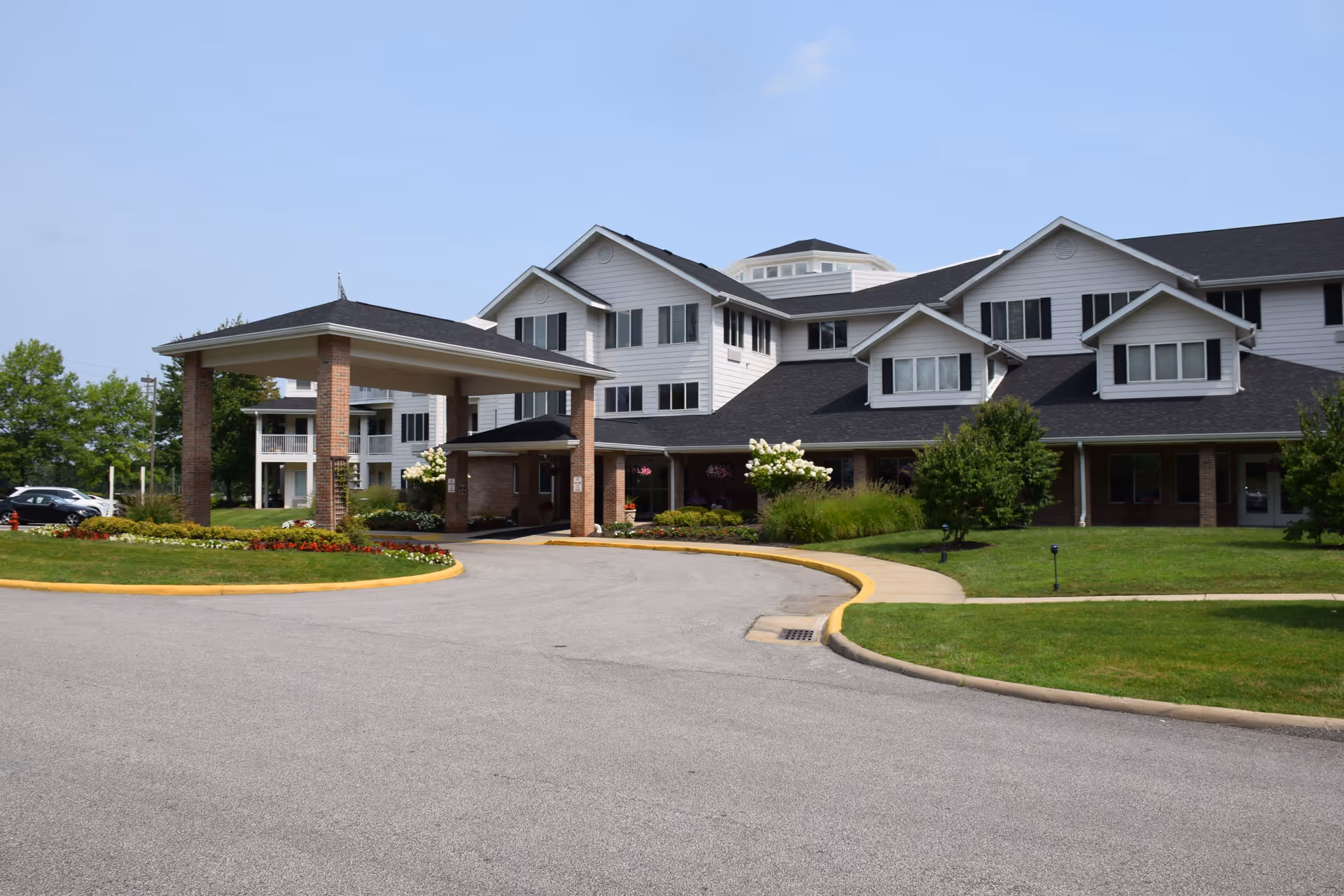 Exterior view of StoryPoint Strongsville senior living facility showing a large, multi-story building with white siding and black shutters. The building has a covered entrance supported by brick columns, surrounded by well-maintained landscaping including green grass, bushes, and flowers. A driveway curves in front of the entrance under a clear blue sky.