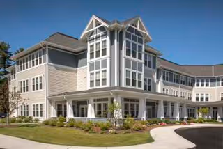 Exterior view of a multi-story senior living facility building with large windows, a covered entrance, landscaped greenery, and a clear blue sky.