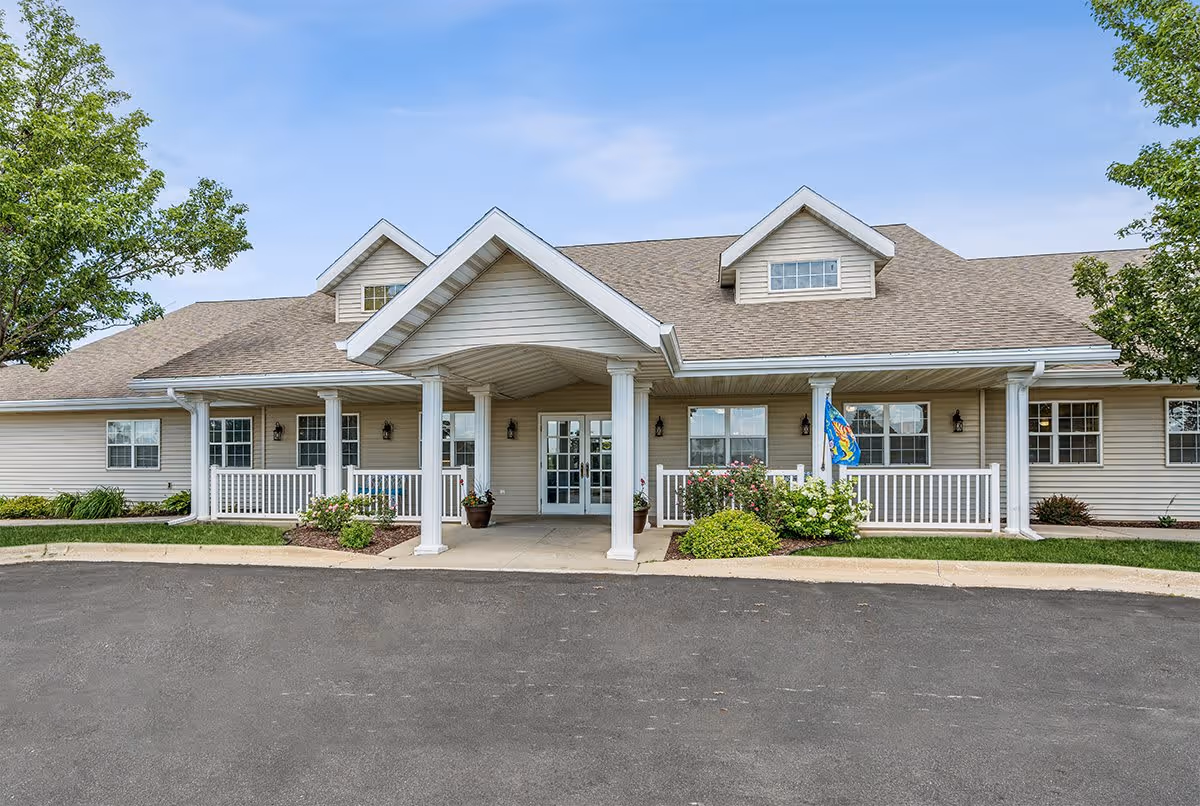 Front exterior view of a single-story senior living facility building with beige siding, a covered entrance supported by white columns, several windows, a small porch with white railings, and landscaping including bushes and trees under a blue sky.