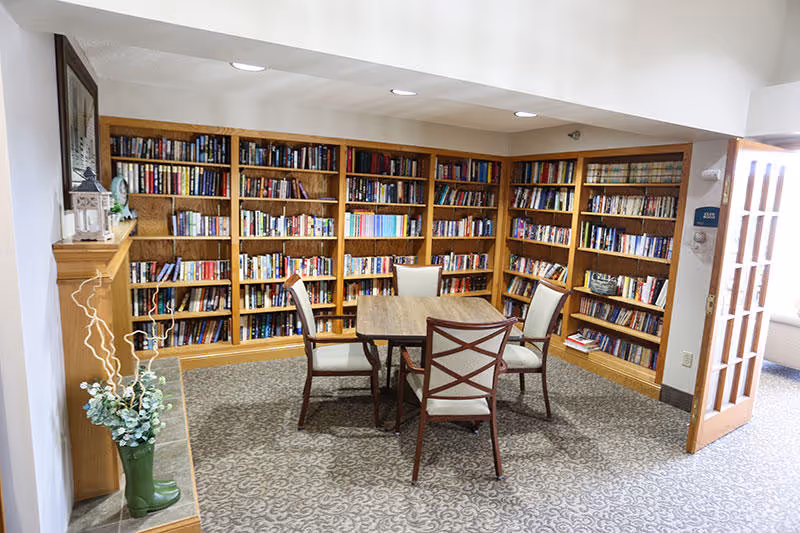 A cozy library room with wooden bookshelves filled with books lining the walls. In the center, there is a wooden table surrounded by four cushioned chairs. The room has a patterned carpet and a wooden mantelpiece with decorative items on the left side. A door with glass panels is open on the right, allowing natural light to enter.