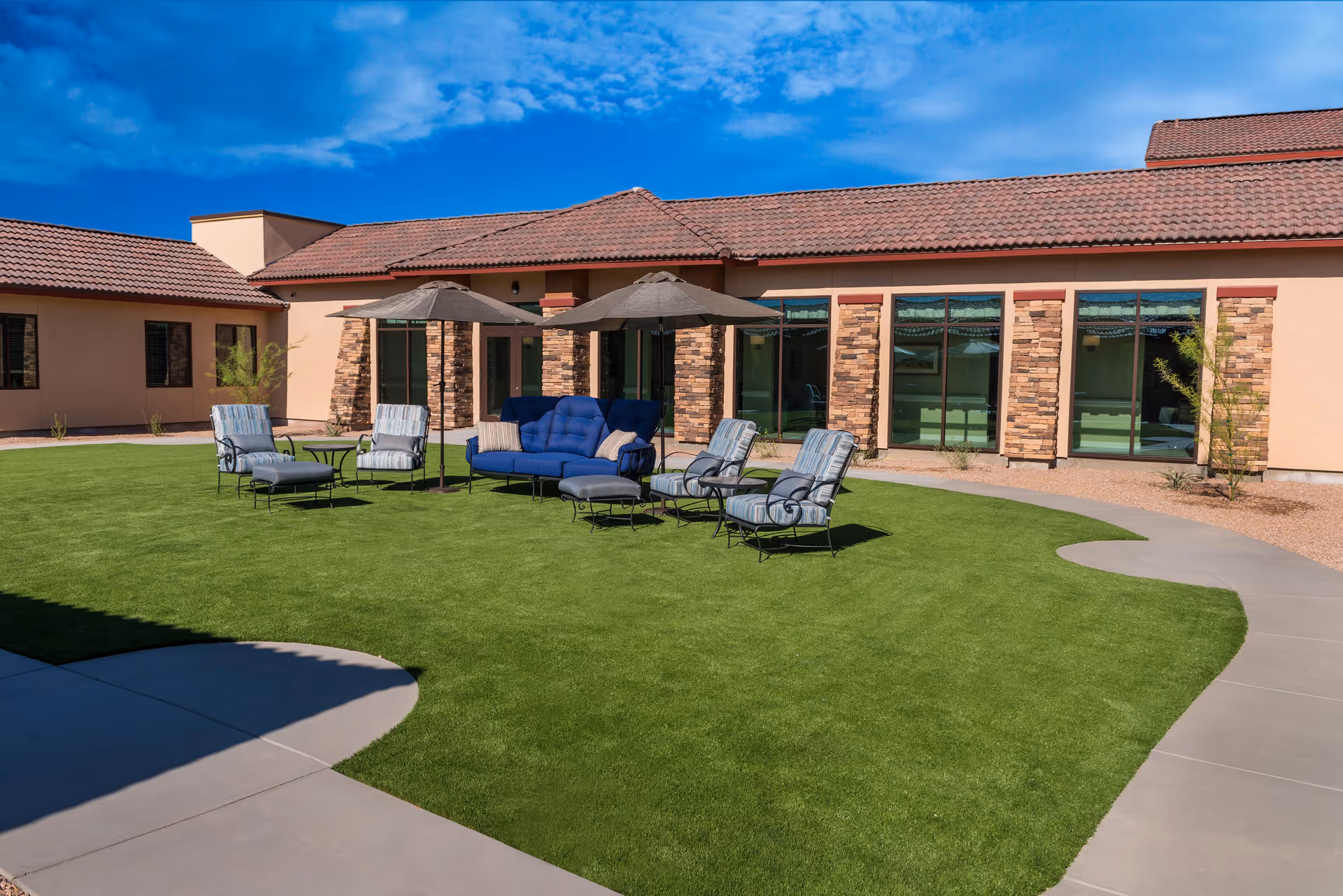 Outdoor courtyard area of Canyon Winds Assisted Living & Memory Care featuring green artificial grass, several cushioned chairs and a sofa with umbrellas providing shade, surrounded by a building with stone and stucco exterior walls under a blue sky.