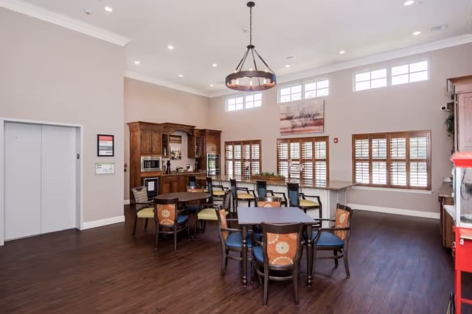 A spacious dining area with multiple tables and chairs, a kitchenette with wooden cabinets, a microwave, and a wine cooler. The room has large windows with wooden shutters, a high ceiling with recessed lighting, and a circular chandelier. There is an elevator door on the left side and a popcorn machine on the right.
