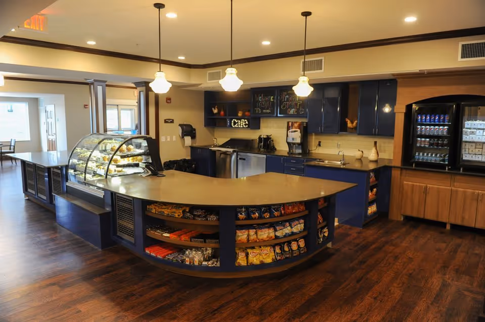 Interior view of a snack and beverage area in a senior living facility. The space features a curved counter with shelves stocked with various chips and snacks. Behind the counter are blue cabinets, a sink, coffee machines, and a refrigerated display case with drinks and other items. The area is well-lit with hanging pendant lights and has wood flooring.