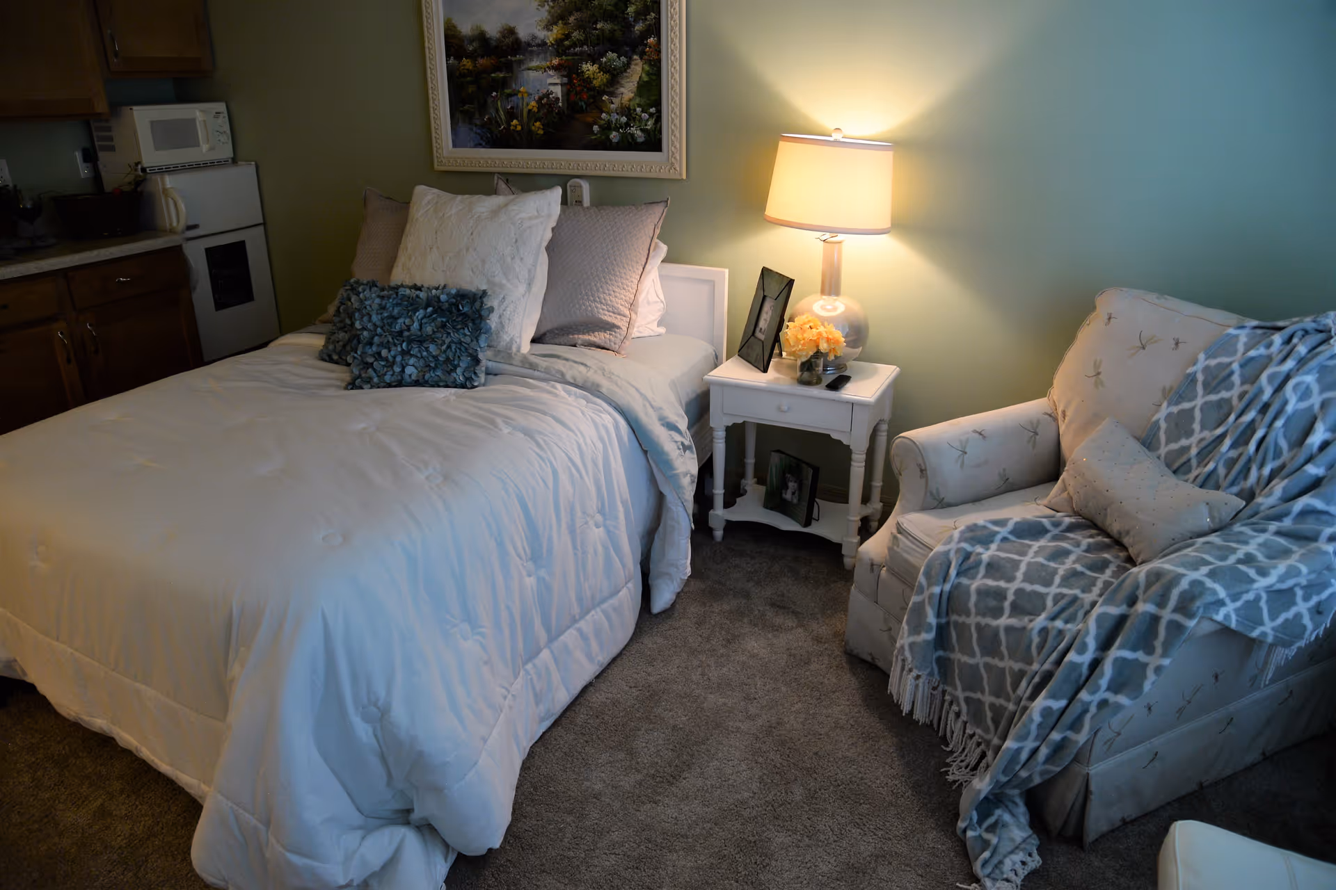 A cozy bedroom with a neatly made bed featuring white and blue pillows and a white comforter. Next to the bed is a white nightstand with a lamp, a small bouquet of flowers, and framed photos. To the right is a cushioned armchair draped with a blue and white patterned throw blanket. The room has carpeted flooring and a painting of a garden scene on the wall above the bed. A small kitchenette with wooden cabinets, a microwave, and a mini fridge is visible in the background.