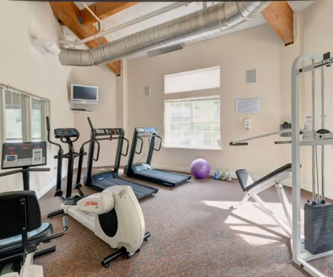 A small fitness room with exercise equipment including two treadmills, an elliptical machine, a stationary bike, a weight machine, and a purple exercise ball near a window. The room has beige walls, a carpeted floor, exposed wooden beams, and a large ventilation duct on the ceiling.