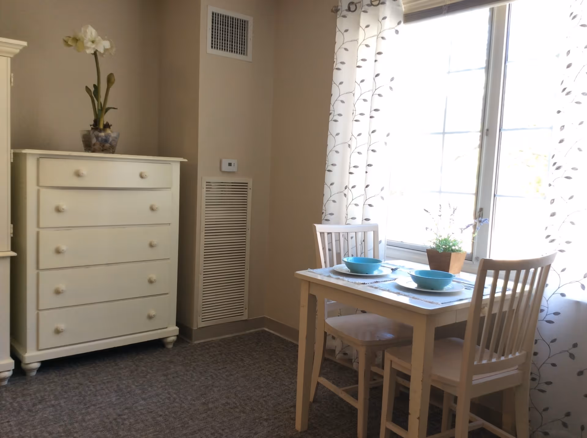 A small dining area with a white wooden table set for two with blue bowls and placemats, next to a large window with white curtains featuring a leaf pattern. To the left, there is a white chest of drawers with a potted plant on top. The room has beige walls and carpeted flooring.