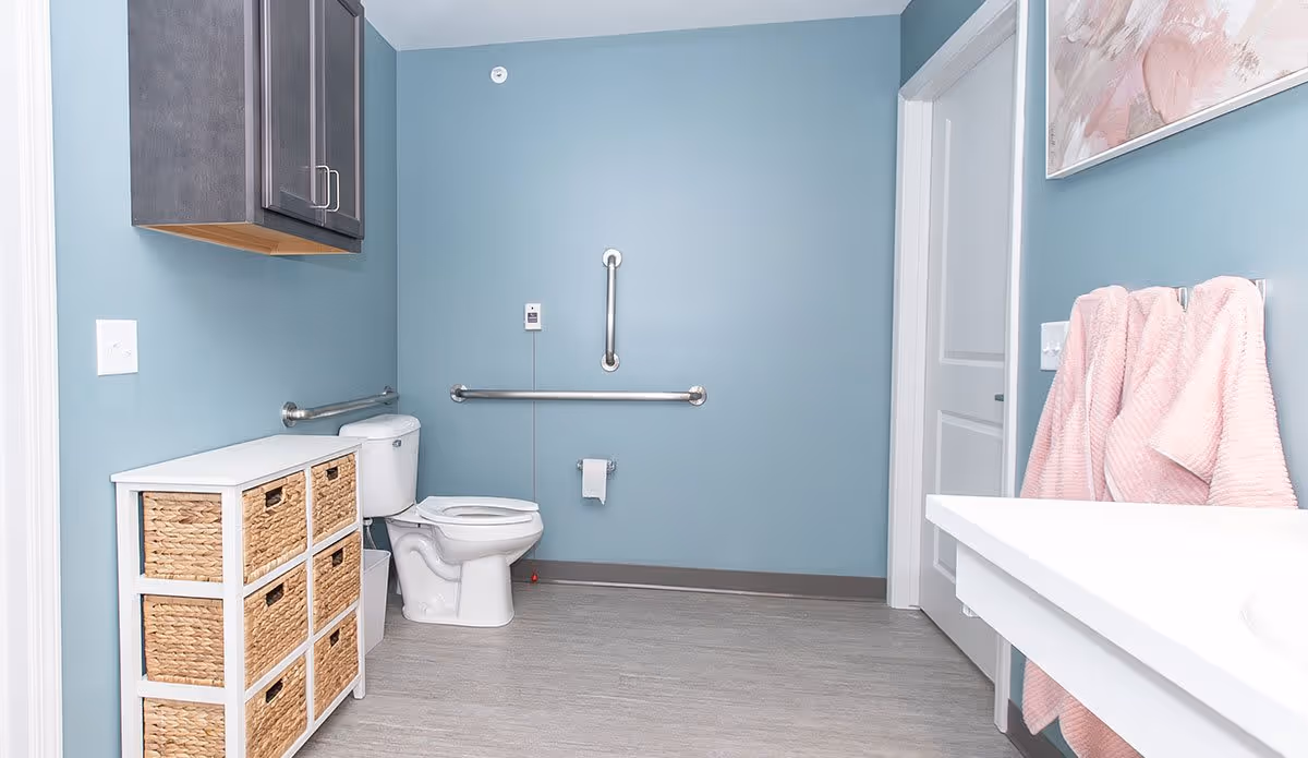 A spacious bathroom with light blue walls and gray flooring. There is a white toilet with stainless steel grab bars on the wall behind and to the side. To the left of the toilet is a white storage unit with six woven baskets. Above the storage unit is a dark gray wall cabinet. On the right side, there is a white countertop with two pink towels hanging on a towel rack beneath a framed floral artwork. A white door is visible in the background.