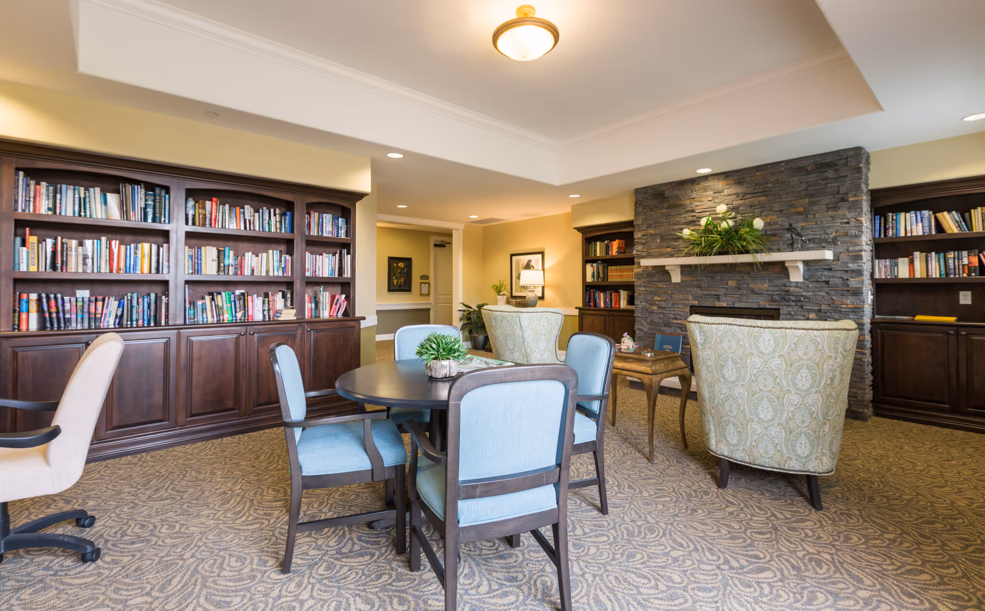A cozy senior living common room with bookshelves, a round table and upholstered chairs facing a stone fireplace.