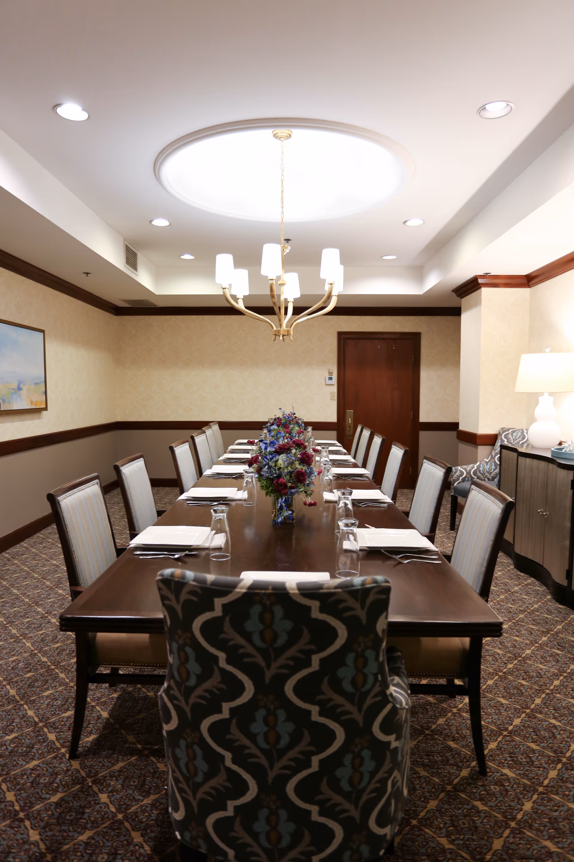 A formal dining room with a long wooden table set for twelve people. The table is decorated with three floral centerpieces and each place setting includes a napkin, fork, knife, and an upside-down glass. The room features patterned carpet, beige walls with a wooden chair rail, a chandelier hanging from a circular ceiling recess, and a sideboard with a lamp and decorative items.