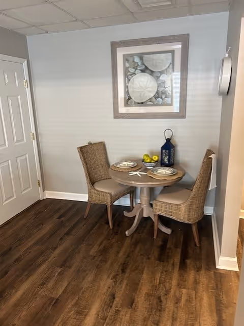 Small dining nook with a round wooden table, two wicker chairs, place settings, a lantern and framed artwork on a pale gray wall.
