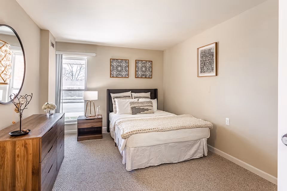 A bright and cozy bedroom featuring a neatly made bed with white bedding and decorative pillows. The room has beige walls adorned with three framed artworks. Next to the bed is a wooden nightstand with a modern lamp and a glass bottle. On the left side, there is a wooden dresser with a round mirror above it and a decorative tree-shaped jewelry holder. A large window with vertical blinds allows natural light to fill the room.