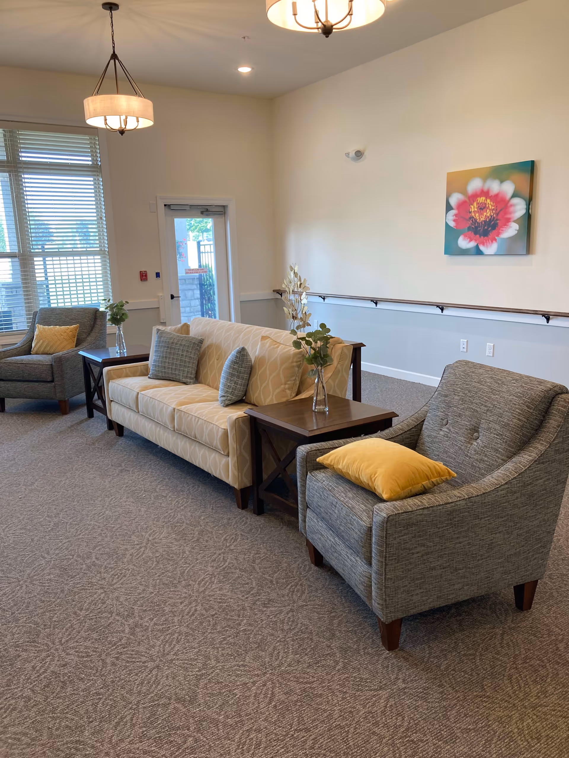 A bright and cozy sitting area with a beige patterned sofa adorned with two gray pillows, flanked by two wooden side tables each holding a glass vase with greenery. There are two gray armchairs, each with a yellow pillow. The room has a large window with blinds, a door leading outside, a floral painting on the wall, and a ceiling light fixture.