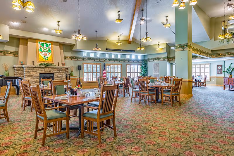 A spacious dining room in a senior living facility with multiple wooden tables and chairs arranged neatly. The room features a patterned carpet, a stone fireplace with a colorful stained glass artwork above it, large windows letting in natural light, and several hanging light fixtures from the ceiling. There are decorative plants and an American flag near the windows.