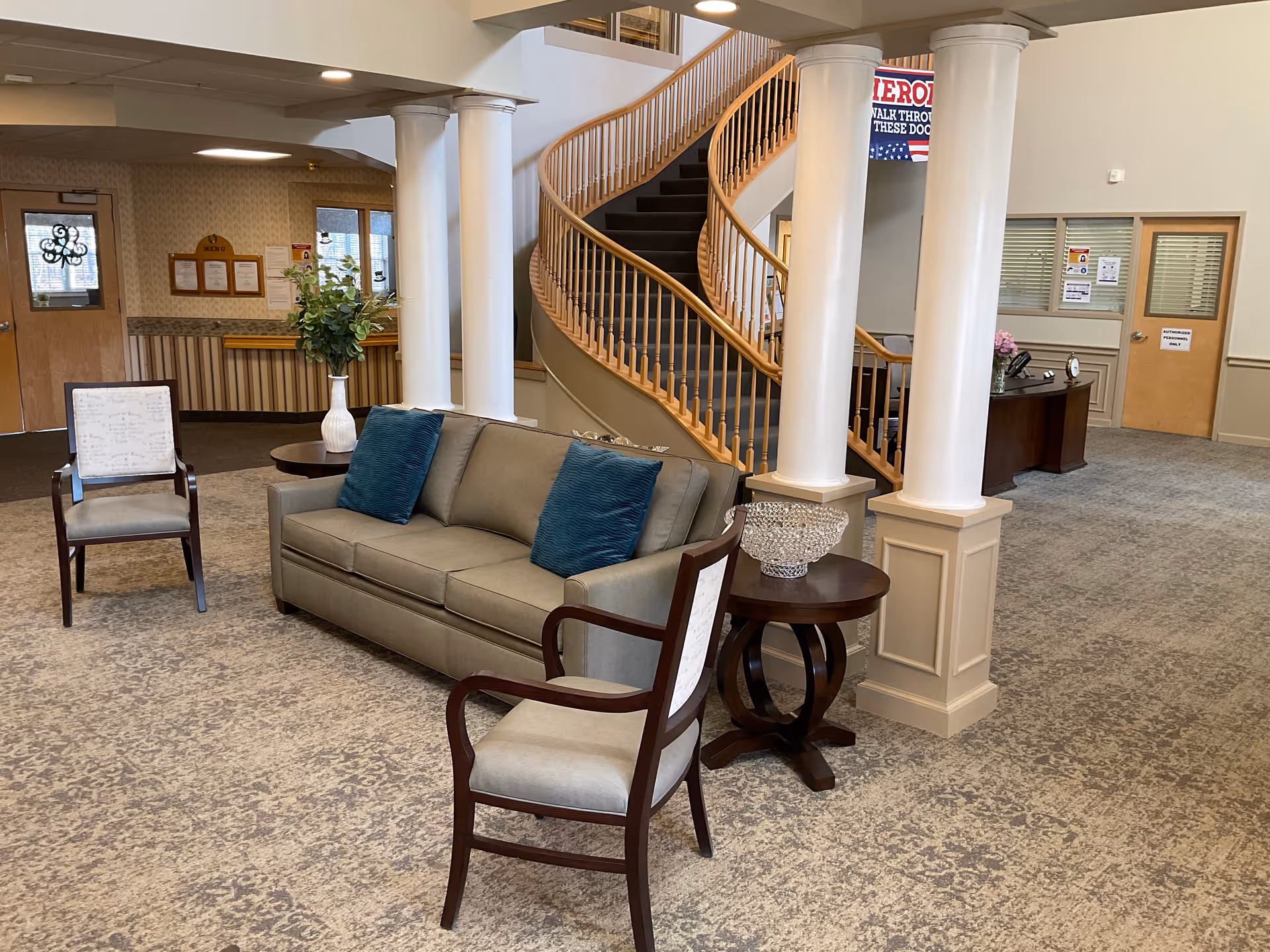 Interior view of a senior living facility lobby area with a gray sofa adorned with two blue pillows, two wooden armchairs with light upholstery, a round wooden side table with a decorative bowl, and white columns. In the background, there is a curved wooden staircase, a reception desk, and doors with signage.