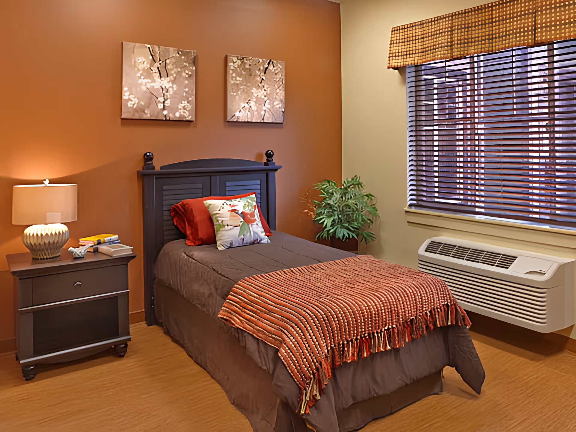 A cozy bedroom with a single bed featuring a dark headboard, brown bedding, a red throw blanket, and decorative pillows. There is a nightstand with a lamp, books, and a tissue box next to the bed. The walls are painted in warm tones with two floral paintings hanging above the bed. A window with wooden blinds and a valance is on the right side, and a green potted plant is placed near the window. Below the window is a wall-mounted air conditioning unit.
