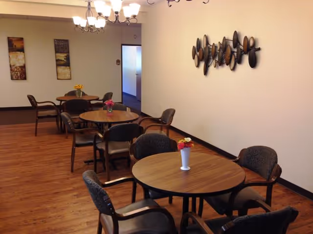 A dining area with round wooden tables and cushioned chairs arranged around them. Each table has a small vase with colorful flowers. The room has wooden flooring, a chandelier hanging from the ceiling, and decorative wall art on the walls.