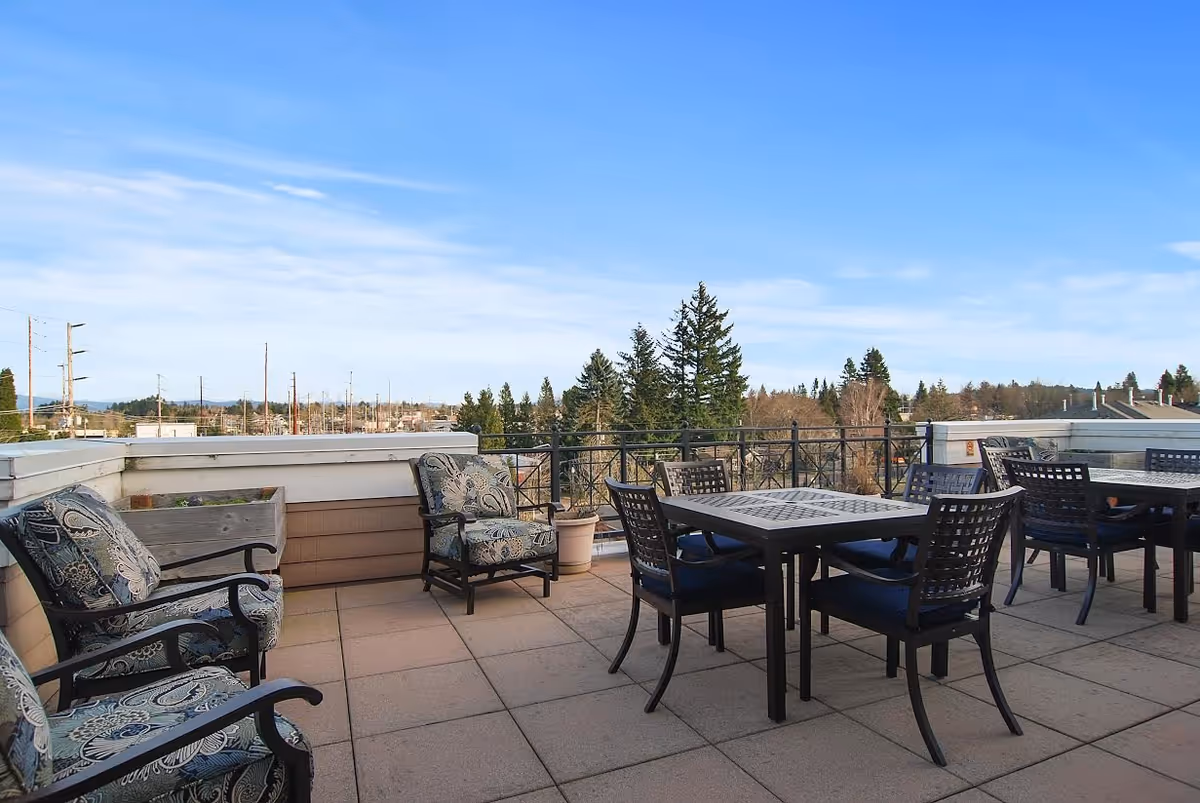 Outdoor patio area with cushioned armchairs and tables with chairs, overlooking trees and a clear blue sky.