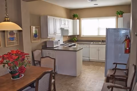 Small communal kitchen and dining area with a table and chairs, white cabinets, refrigerator, and potted plants under a large window.