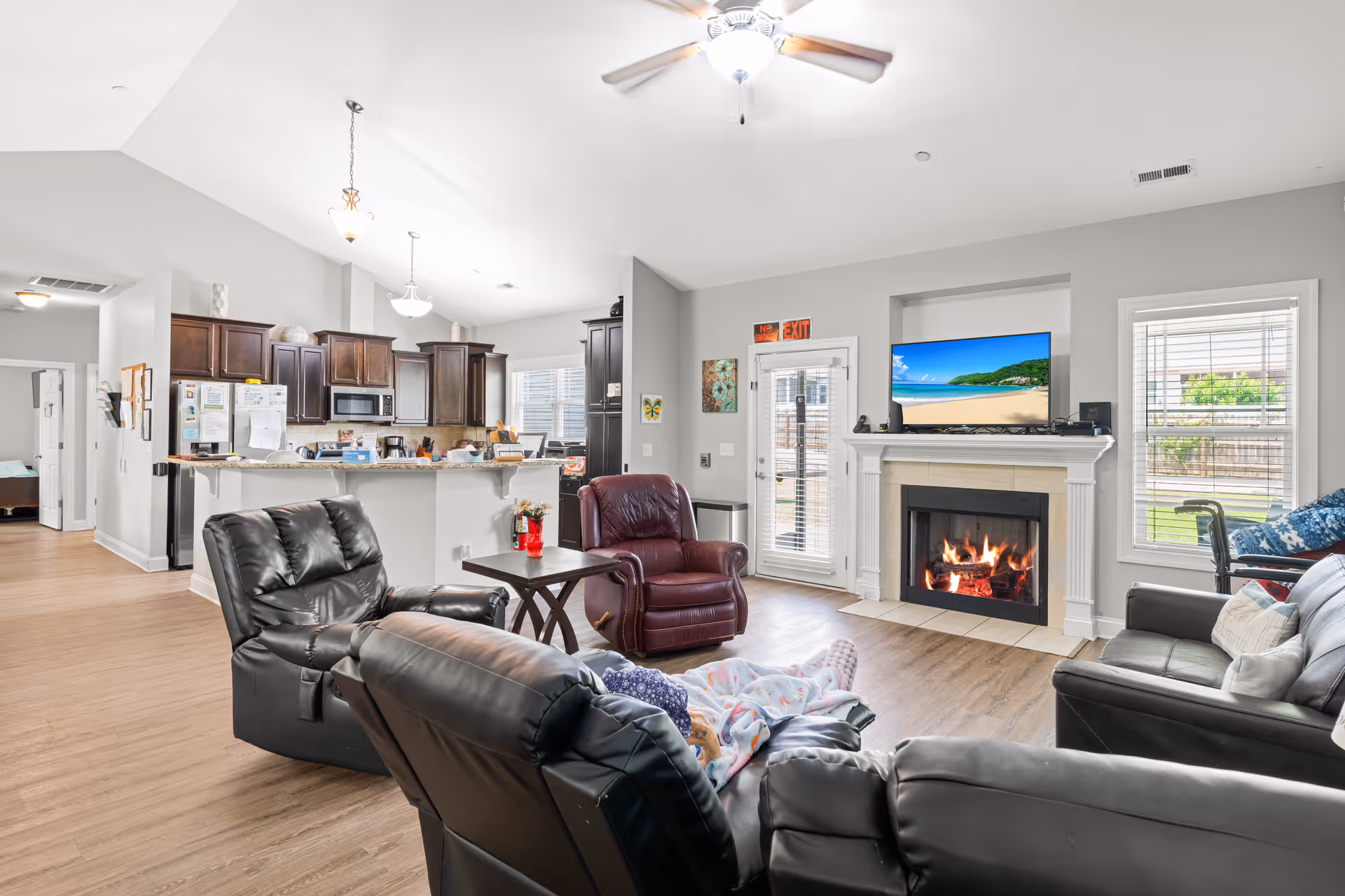 Bright common living area with leather recliners arranged around a fireplace and TV, open to a kitchen with dark cabinets.