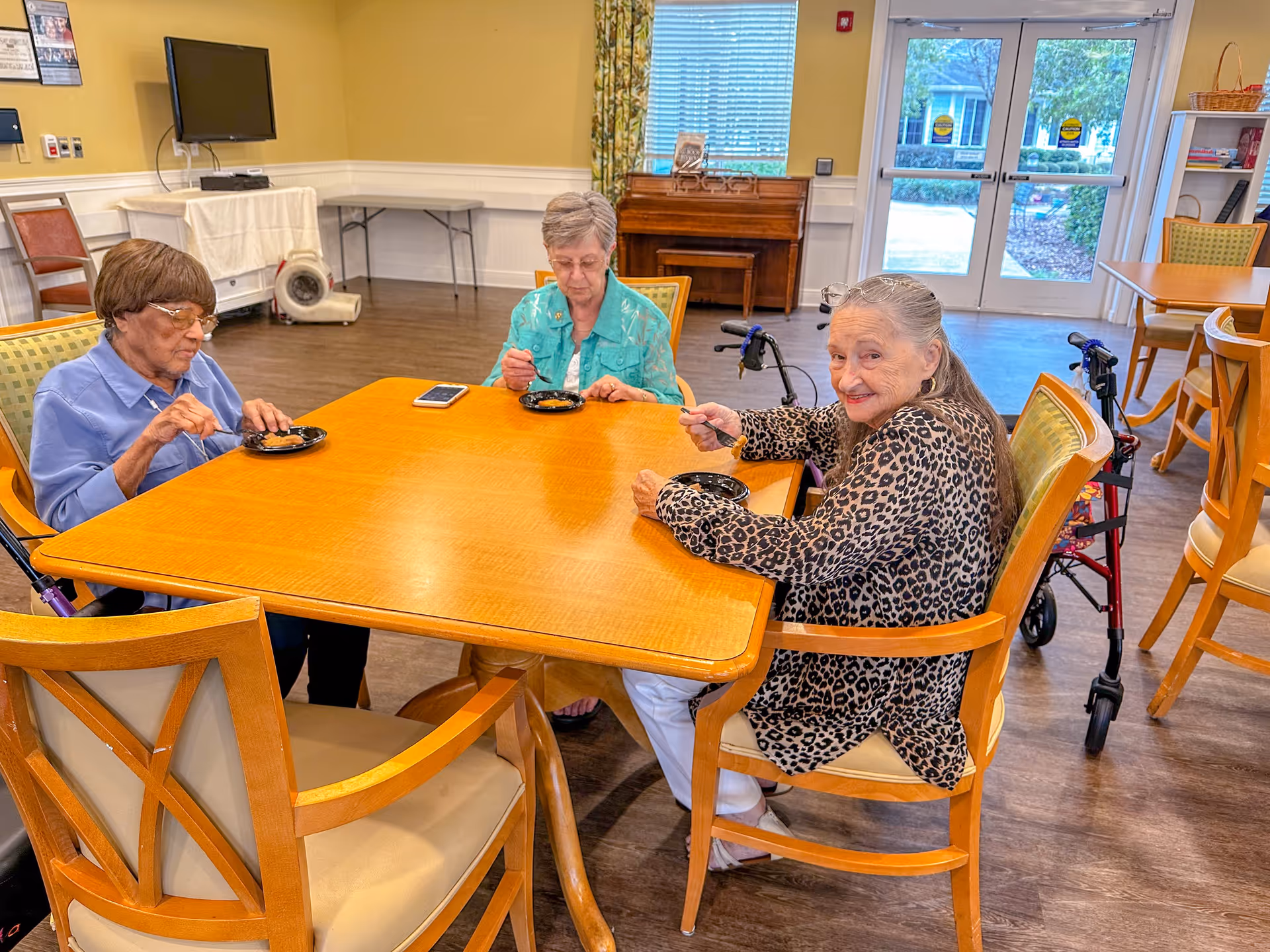 Three senior women sit around a wooden table eating dessert in a bright communal dining room.