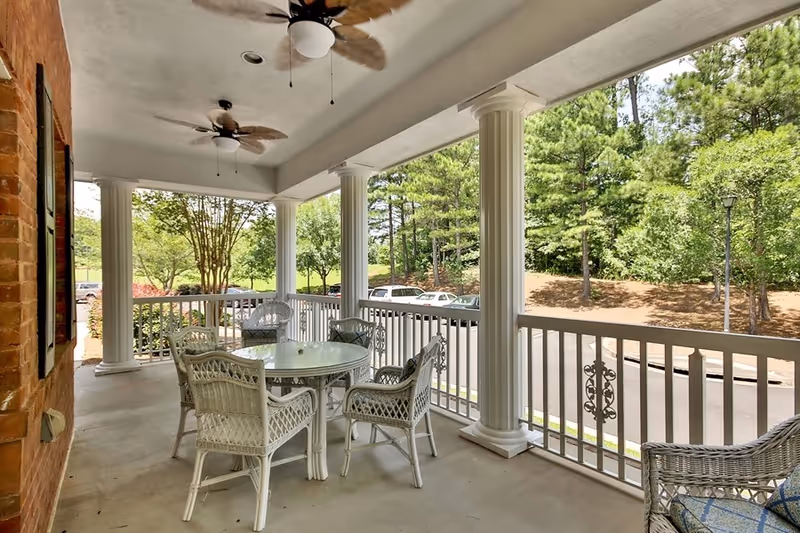 Covered outdoor patio area with white wicker chairs and a round glass table. The patio has white columns and ceiling fans with leaf-shaped blades. Beyond the railing, there is a view of a parking lot with cars and green trees in the background.