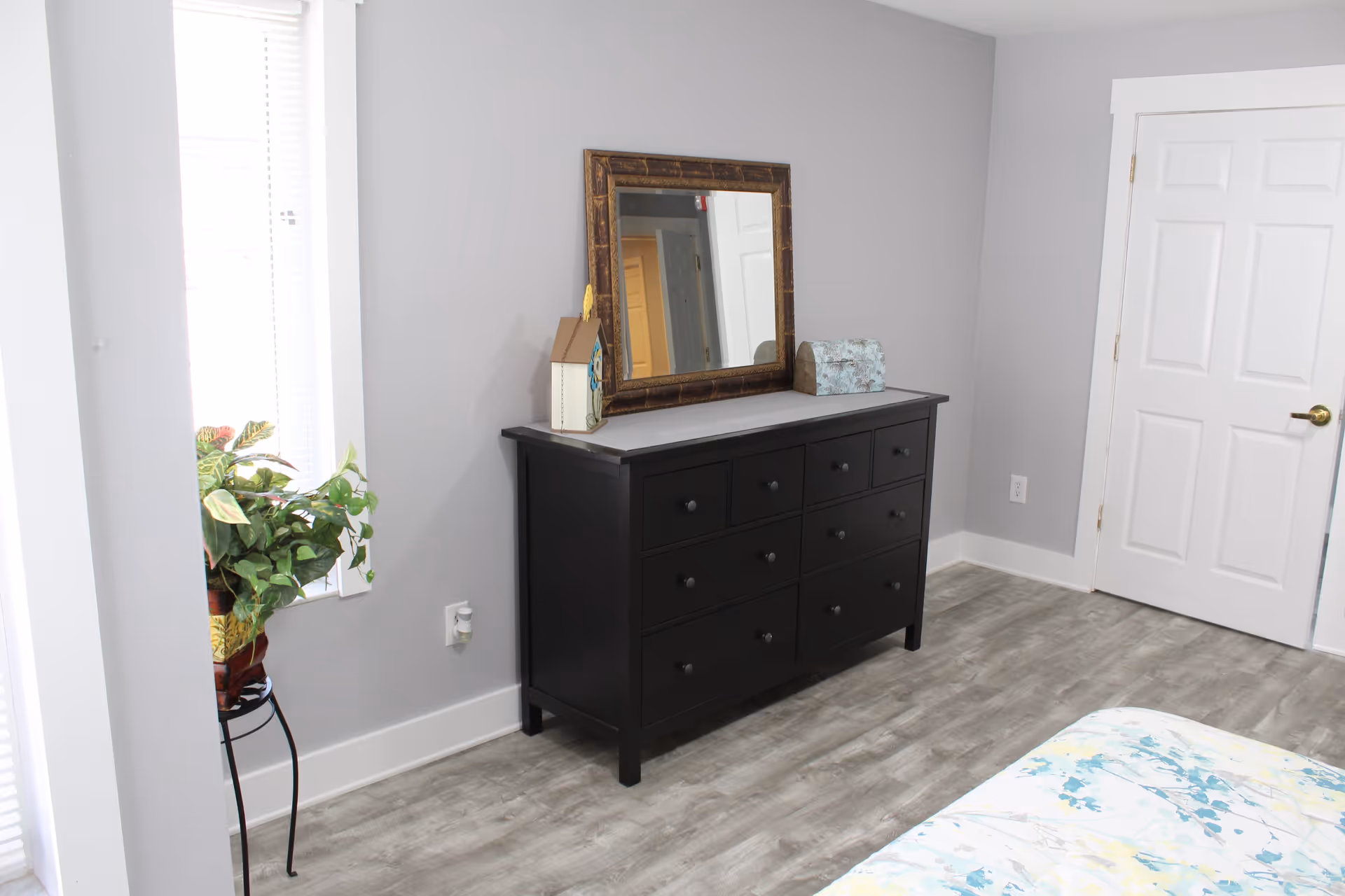 A bedroom corner with a black dresser featuring multiple drawers, a large framed mirror resting on top, and decorative items including a small house-shaped ornament and a box. To the left, there is a window with blinds and a potted plant on a stand. The floor is light wood, and a white door with a gold handle is visible on the right side.