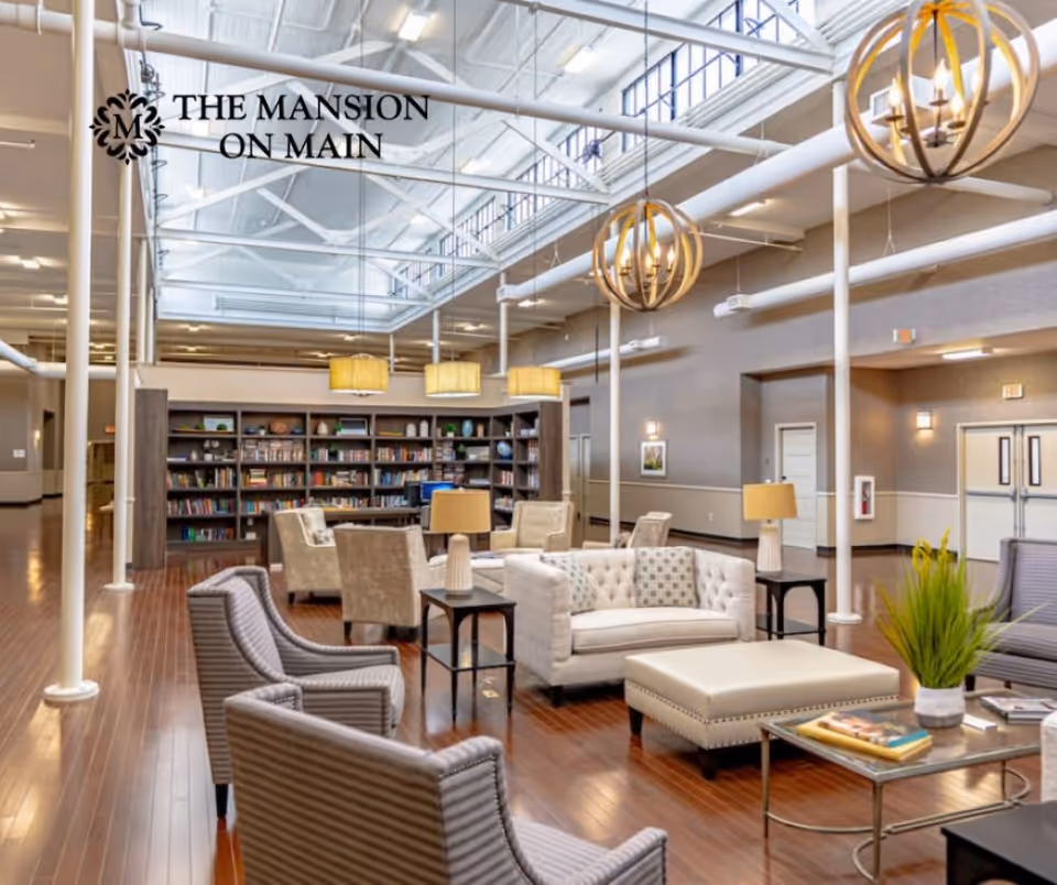 A spacious and well-lit common area in a senior living facility featuring comfortable seating including armchairs, a tufted sofa, and ottomans arranged around coffee tables. The room has wooden flooring, modern hanging light fixtures, and a large bookshelf filled with books and decorative items along the back wall. The ceiling is high with exposed beams and skylights allowing natural light to fill the space.