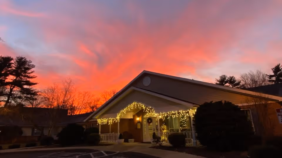 Exterior view of a single-story building at sunset with a vibrant orange and pink sky. The building is decorated with string lights along the roofline and has a small porch area with a wreath on the door. Trees and bushes surround the building.