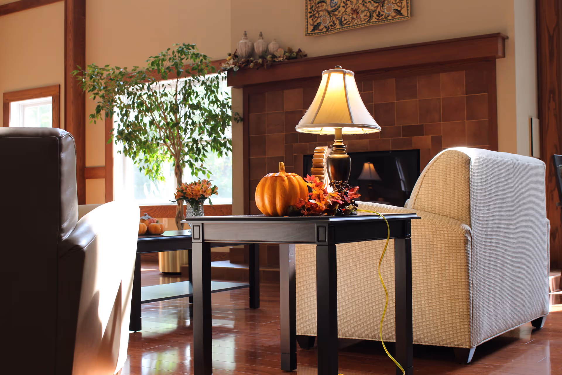 Cozy living room with armchairs and a side table topped by a lamp and fall decorations in front of a fireplace.